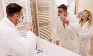 Man applies shaving cream with a safety razor in the mirror, woman smiles nearby.
