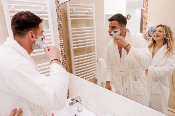 Man applies shaving cream with a safety razor in the mirror, woman smiles nearby.