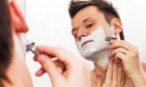 Man shaves face with a safety razor and shaving cream in mirror.