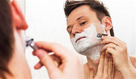 Man shaves face with a safety razor and shaving cream in mirror.
